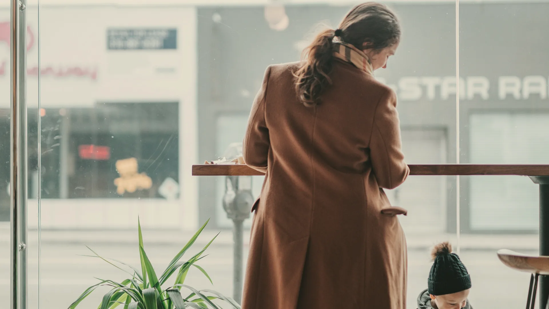 A mother at a café window counter while her toddler plays nearby